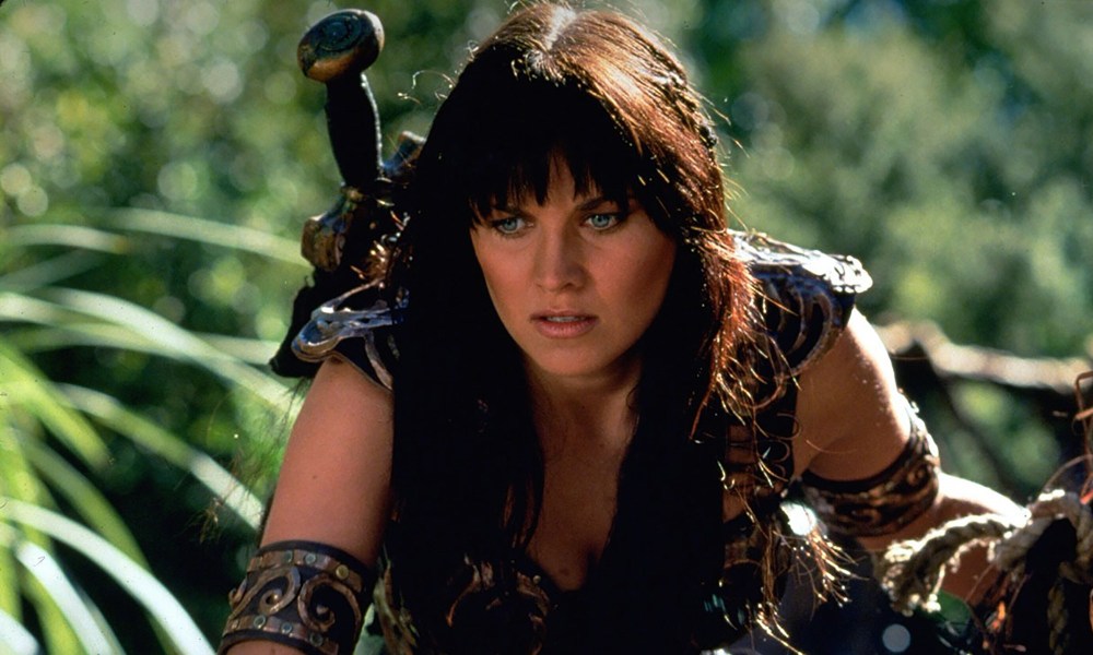 Xena crouched down in the grass with weapons on her back in Xena: Warrior Princess.